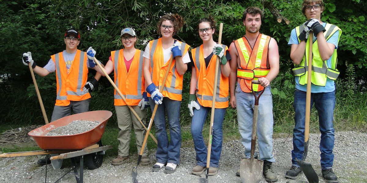 Row of young trail workers wearing orange safety vests and holding shovels and rakes